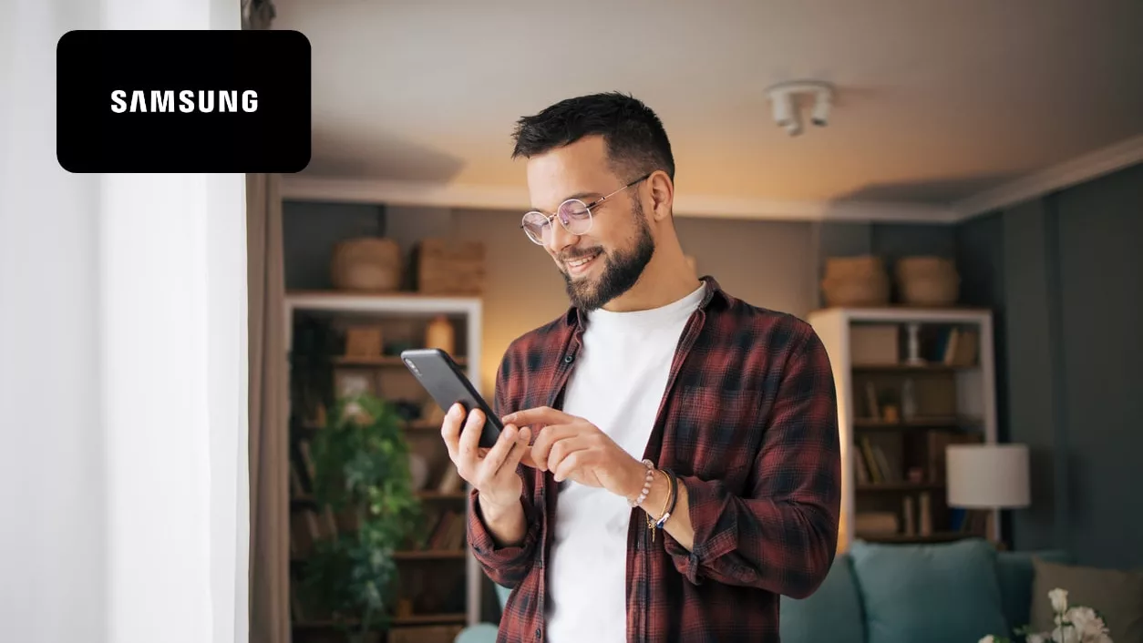 An adult man is standing in a modern living room by the window and using a smartphone to talk.