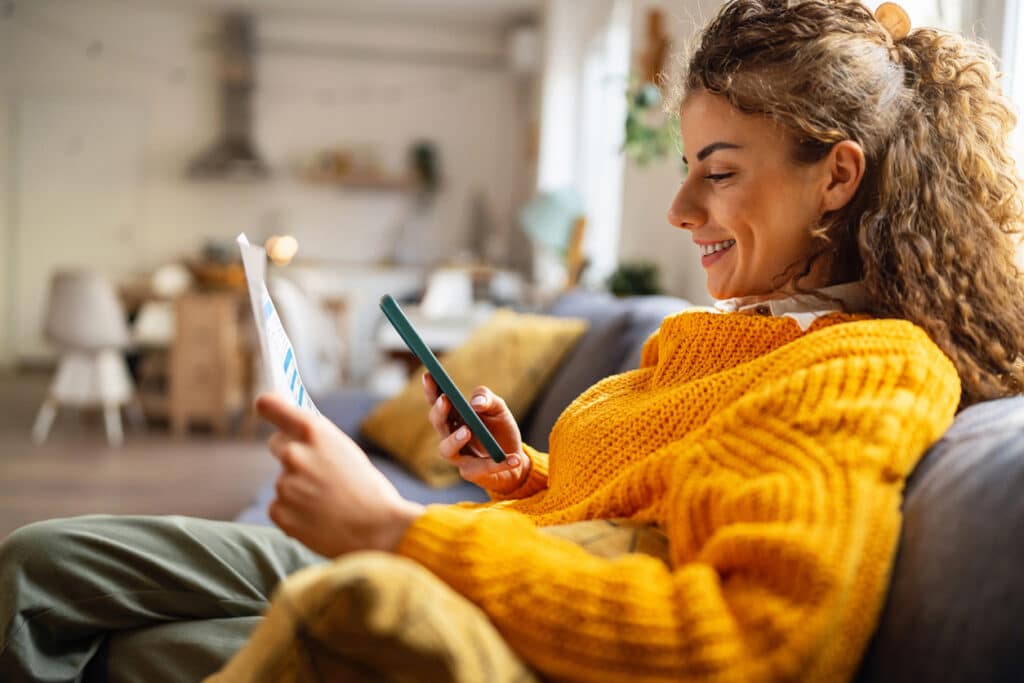 Frau in gelbem Pullover checkt Bankauszüge auf der Couch