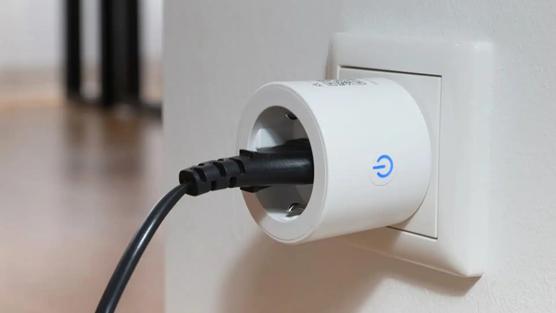 Weiße Steckdose in der ein runder weißer Stecker steckt in dem ein schwarzer Gerätestecker mit Kabel steckt an weißer Wandecke