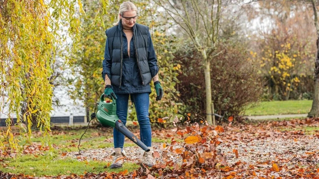 Frau in dunkler Weste und Jeans mit Schutzbrille und grünem Bosch-Laubbläser auf Wise mit Laub vor Bäumen mit gelben Blättern