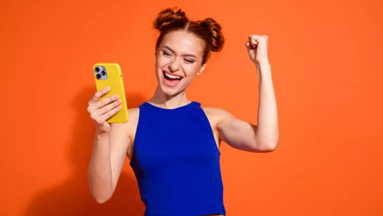 regte junge Frau in stilvollem Freizeitoutfit, die über Handy vor orangefarbenem Hintergrund feiert