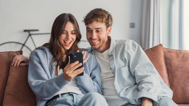 Junges Paar sitzt auf dem Sofa und schaut auf ein Handy.