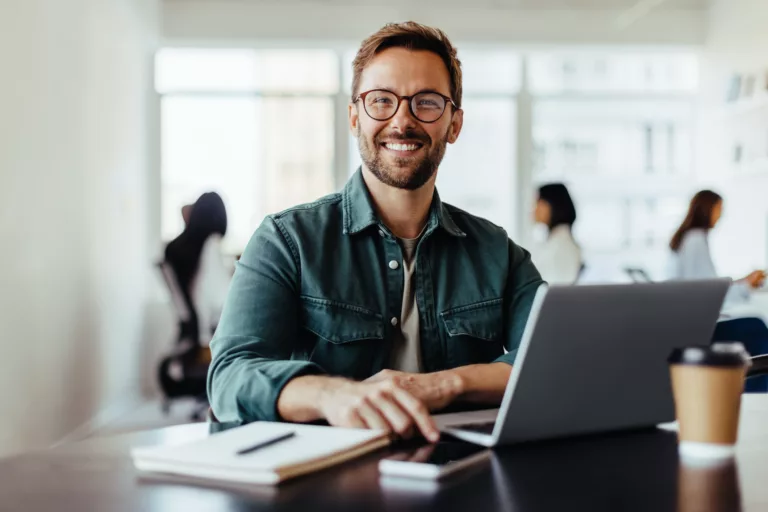 Fröhlicher Mann grinst in die Kamera, er sitzt in einem Büro an einem Laptop (@istockphoto.com)