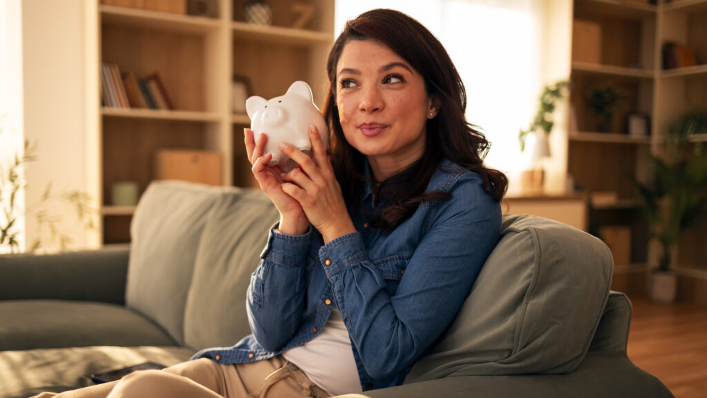 Junge Frau sitzt auf dem Sofa mit einem Sparschwein in der Hand.