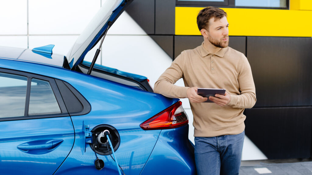 Ein junger Mann lädt ein Elektroauto auf, während er an einer Station steht und ein Tablet in der Hand hält – Stockfoto