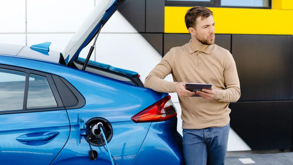 Ein junger Mann lädt ein Elektroauto auf, während er an einer Station steht und ein Tablet in der Hand hält – Stockfoto