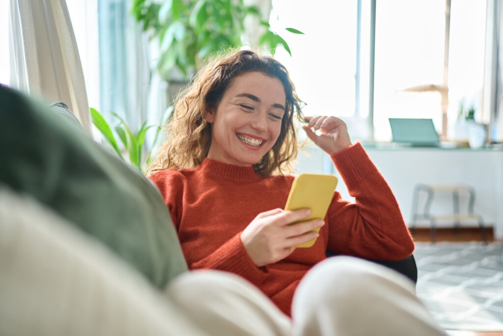 Happy relaxed young woman sitting on couch using mobile phone technology.