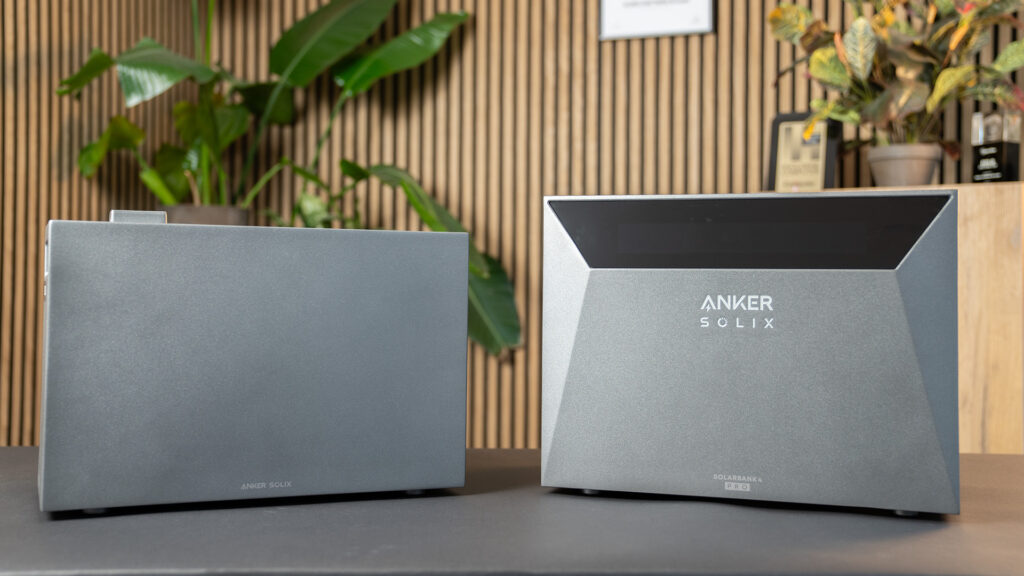 Die graue Anker Solix Solarbank 4 rechts mit Erweiterungsakku links vor braunem Holz mit grünen Pflanzen (@Anker)