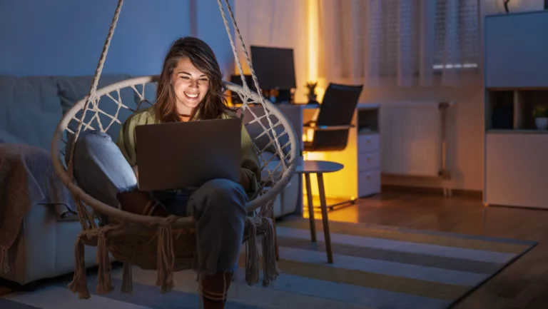 Frau genießt ihre Freizeit zu Hause, sitzt in einem Hängesessel im Wohnzimmer, nutzt einen Laptop, chattet online mit Freunden und entspannt spät in der Nacht. (@istockphoto)