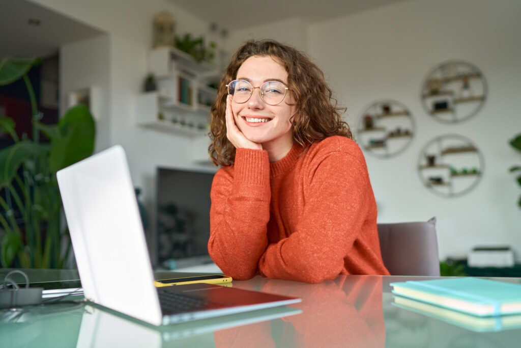 Junge, hübsche, lächelnde Geschäftsfrau beziehungsweise Studentin sitzt am Tisch im Homeoffice mit Laptop, blickt in die Kamera. (@istockphoto)