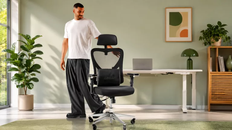 Dunkelhaariger junger Mann mit Bart in weißem Tshirt und schwarzer weiter Hose lächelt und fässt einen schwarzen Bürostuhl, der vor ihm steht an. Hinterihm ein weißer moderner Schreibtisch in einem modernen Zimmer