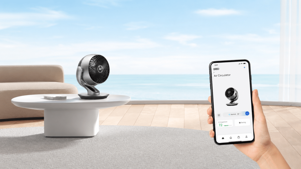 Silberner Tischventilator schräg von der Seite auf einem weißen runden Tisch vor einem beigen Sofa draußen auf einer Lounge mit mehr und blauem Himmel im Hintergrund, Hand hält Smartphone rechts ins Bild, worauf App-Steuerung zu sehen ist