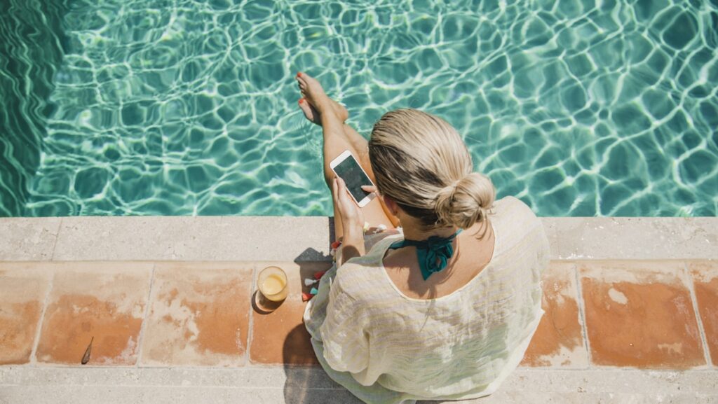 Blonde Frau von oben sitzt am Poolrand mit Beine im Wasser und bedient weißes Smartphone
