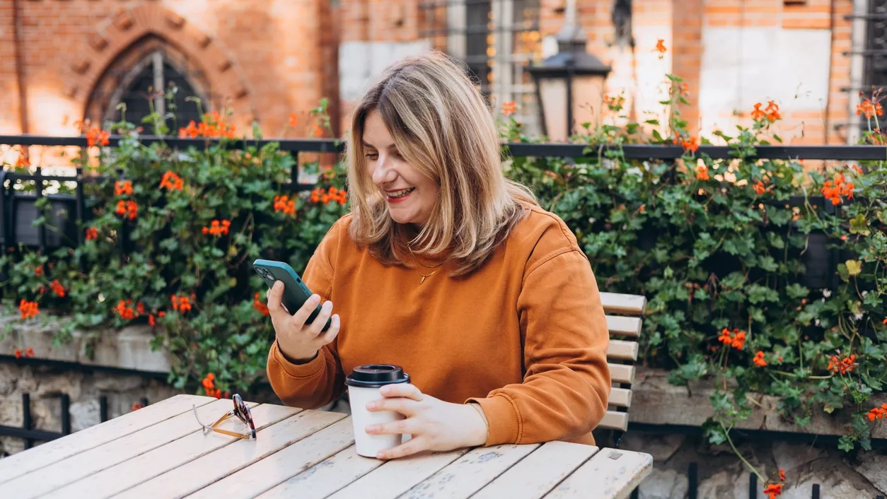 Blonde Frau in orangenem Sweatpulli an Holztisch mit Kaffee-To-Go-Becher und Smartphone in den Händen, lächelt, vor Zaun mit Gewächs mit orangenen Blüten, im Hintergrund Backsteingebäude