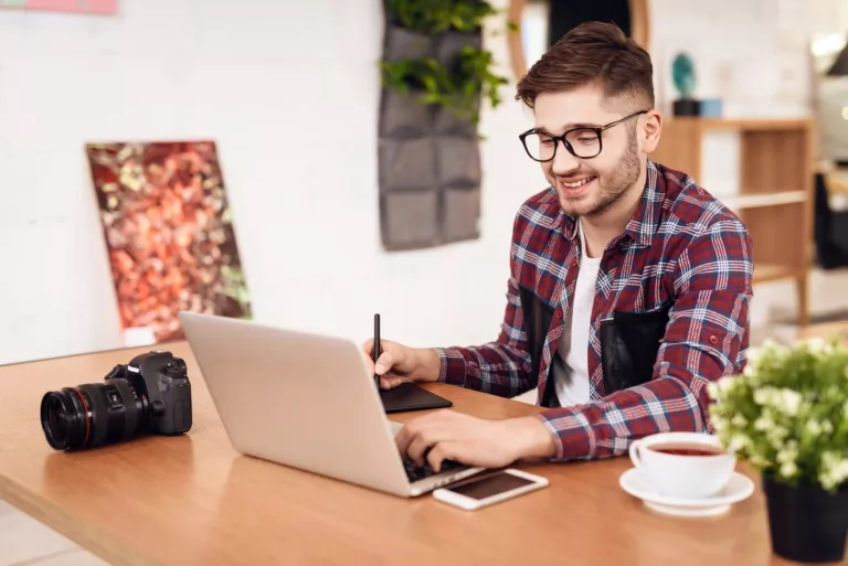 Selbstständiger Fotograf arbeitet am Laptop und schreibt auf einem Tablet im Homeoffice, mit Kaffee und Fotokamera auf dem Schreibtisch.