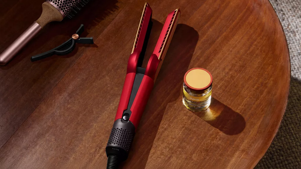 Rot goldenes Glätteeisen von Dyson leicht geöffnet auf dunklem runden Holztisch, daneben rosa Lockenbürste und gelb goldenes Gläschen mit Creme