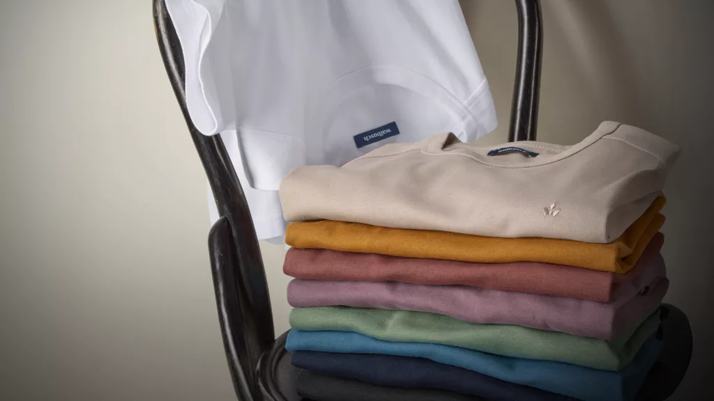 Ein Stapel Walbusch-T-Shirts in gedeckten Farben liegt auf einem Stuhl