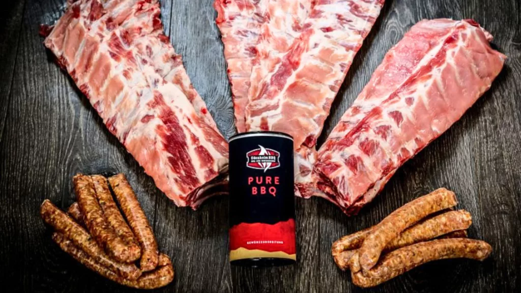 Einige Rippchenleitern und Salsiccia-Würste liegen auf einem Tisch, in der Mitte der Pure BBQ Rub.