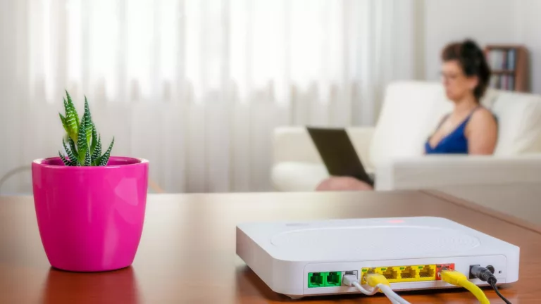 Eine Person sitzt auf dem Sofa und schaut auf ein Tablet, im Vordergrund ein Tisch mit einem W-LAN-Router neben einer Pflanze (@istock)