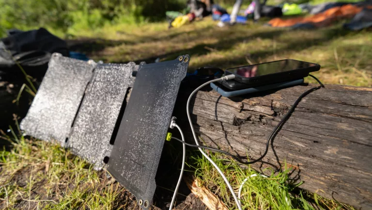 Handys laden an einem Solarpanel im Wald, im Hintergrund sind Zelte zu sehen.