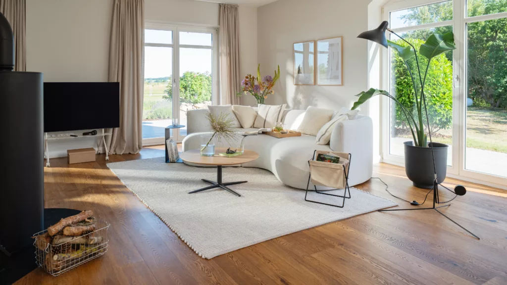 Heller freundlicher Wohnbereich mit weißen Wänden und hellem Holzboden weißem Sofa auf weißem Teppich und bodentiefe Fensterfront an der Seite mit Blick ins Grüne