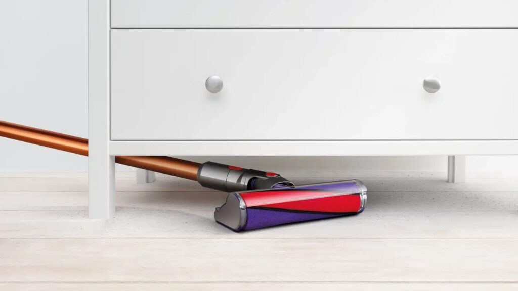 Unterer Teil einer weißen Kommode unter der ein Dyson-Akkusauger mit Kupfer farbenem Stiel und Lila-rotem Aufsatz geschoben wird auf hellem Holzboden