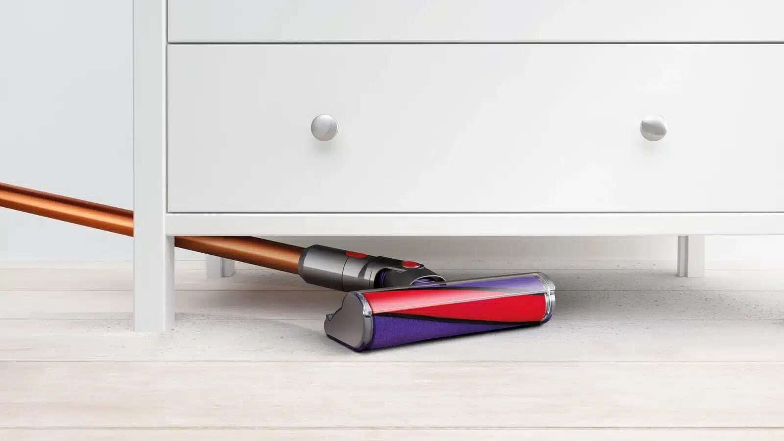 Unterer Teil einer weißen Kommode unter der ein Dyson-Akkusauger mit Kupfer farbenem Stiel und Lila-rotem Aufsatz geschoben wird auf hellem Holzboden