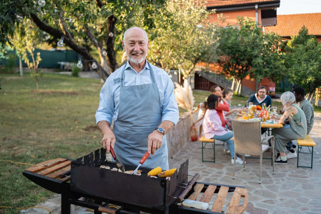 Älterer Mann steht lächelnd am Grill, seine Familie sitzt im Hintergrund am Gartentisch.