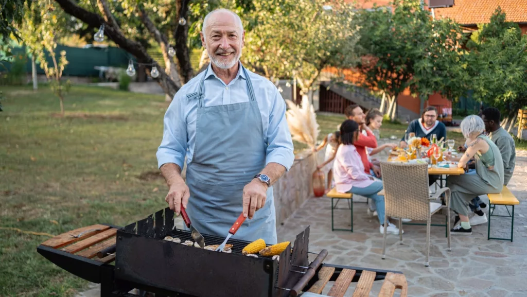 Älterer Mann steht lächelnd am Grill, seine Familie sitzt im Hintergrund am Gartentisch.