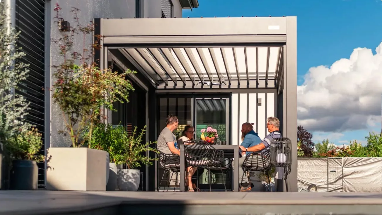 Vier unterschiedliche Menschen sitzen an Tisch unter einer großen grauen Lamellen-Pergola an grauer Hauswand mit Pflanzenkästen drum herum und rechts blauer Himmel