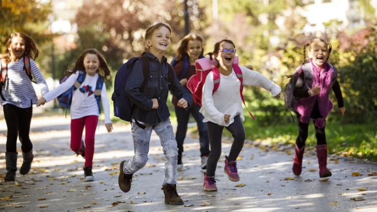 5 Mädchen und ein Junge rennen lachend mit Schulranzen über Fußweg mit etwas Laub. Bäume im Hintergrund