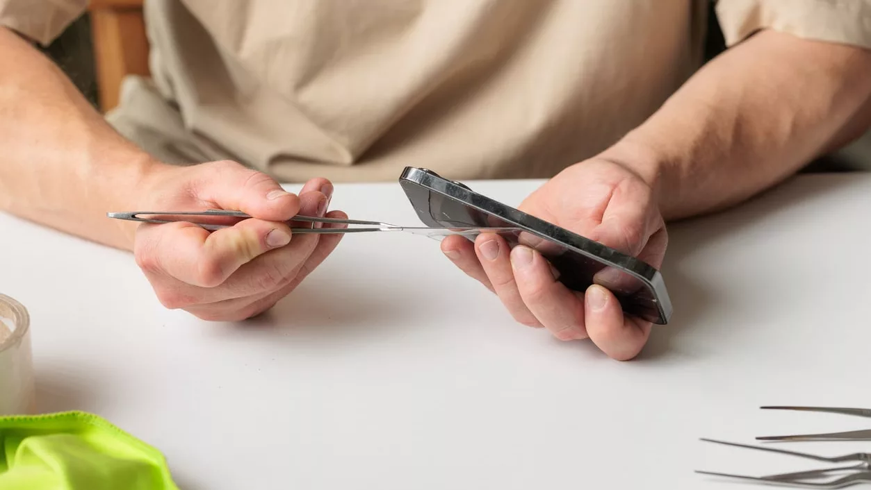 Nahaufnahme von Händen, die im Rahmen eines kleinen Reparaturdienstes zu Hause eine Handy-Displayschutzfolie austauschen.