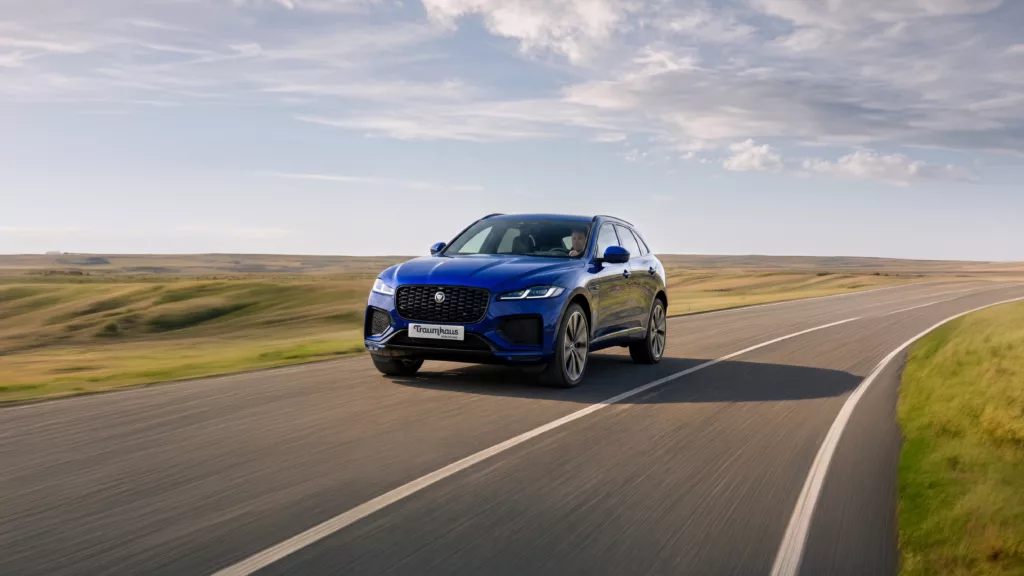 Blauer Jaguar SUV fährt Kurve auf Landstraße von vorne, grüne verschwommene Wiesen an den Seiten, blauer Himmel mit leichten Wolken