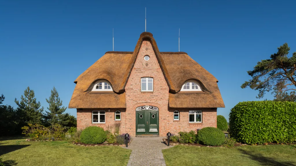 Haus mit spitzem Reetdach von vorne, Fußweg zwischen Rasen, blauer Himmel
