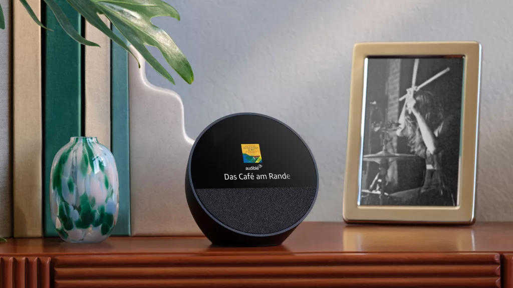 Schwarzer runder Amazon Smartspeaker zeigt Hörbuch Cover und Titel an auf dunkler, glänzender Holzkommode, rechts ein goldener Bilderrahmen hochkant mit schwarz weiß Bild von Drummer, links eine grün weiße Dekovase und ein reinragendes Pflanzenblatt vor grauer Wand