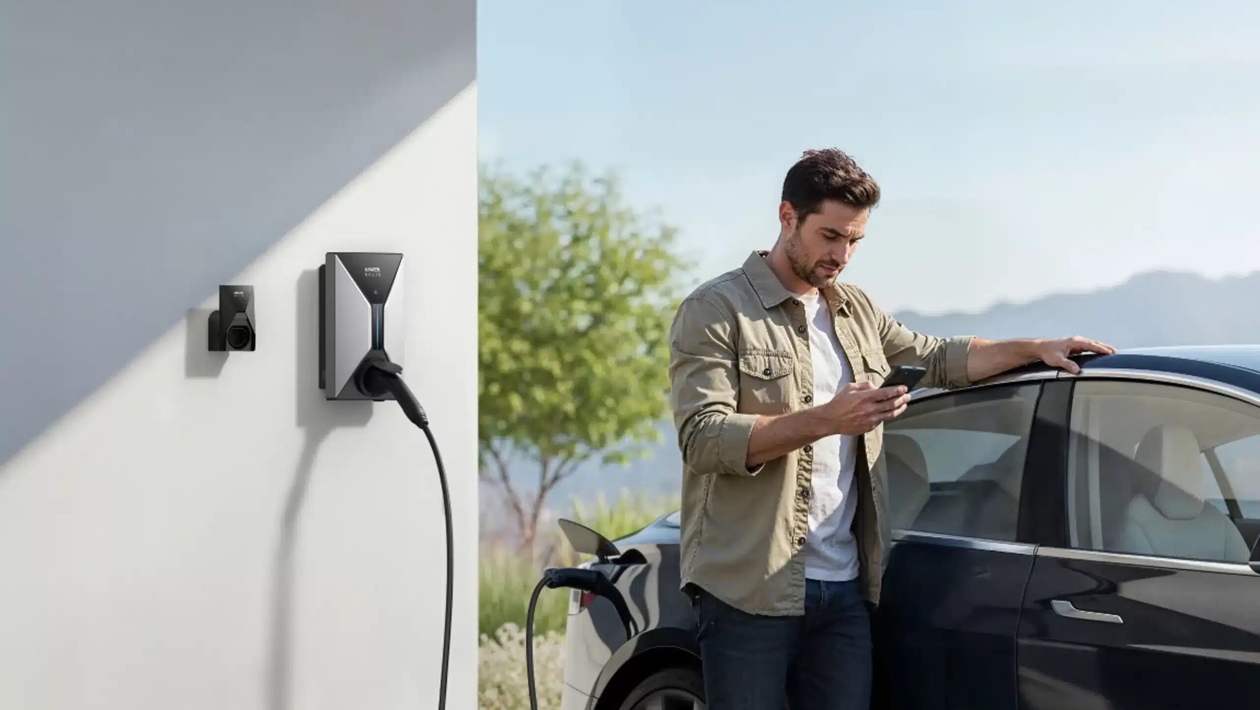 Junger, dunkelhaariger Mann mit Bart steht seitlich an schwarzem Auto und guckt auf Smartphone, links neben ihm eine weiße Hauswand an der eine rechteckige silber schwarze Wallbox hängt und ein schwarzes Kabel verbindet Box und Auto. Blauer Himmel und Bergpanorama im Hintergrund