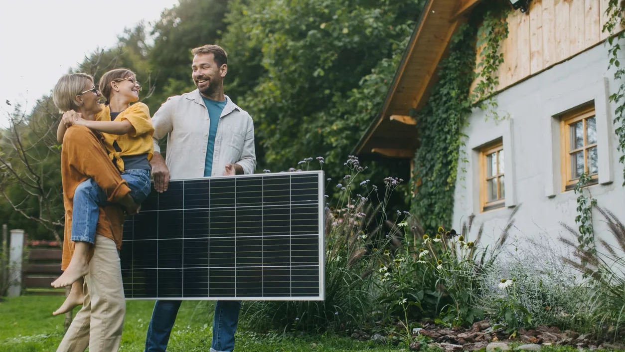Links blonde Frau mit hellbraun haarigem Mädchen auf dem Arm, daneben ein braunhaariger Mann, der ein schwarzes Solarmodul waagerecht hält, alle lächeln vor grünem Garten, rechts Ecke von weißem Haus mit Holzverkleidung
