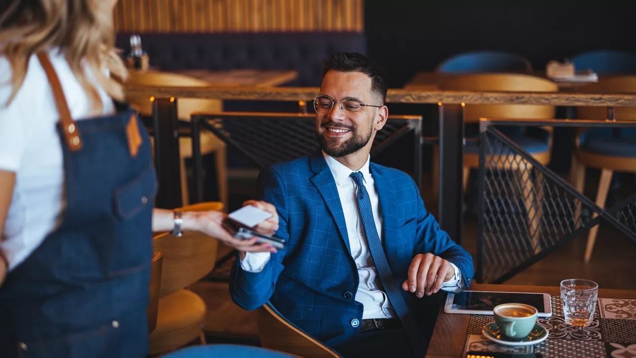 A smiling businessman in a suit pays for coffee using a credit card in a contemporary cafe setting, showcasing modern dining and payment methods.