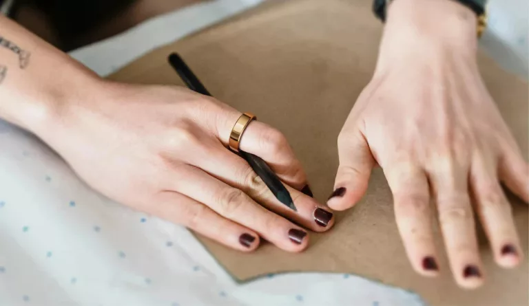 Zwei feine Hände mit schwarz lackierten Nägeln ziehen Pappe glatt. Rechte Hand hält schwarzen Stift und hat einen goldenen Ring am Zeigefinger