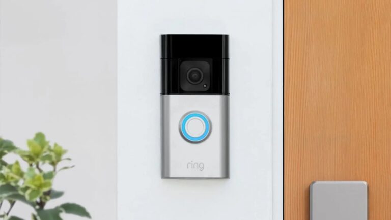 Silbern schwarze Ring-Videotürklingel rechteckig hochkant auf weißer Wand neben heller Holztür und grüner Pflanze links