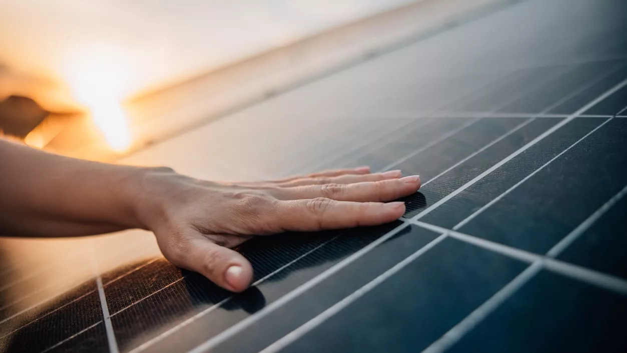 Hand von links berührt dunkles Solarpanel bei Sonnenuntergang