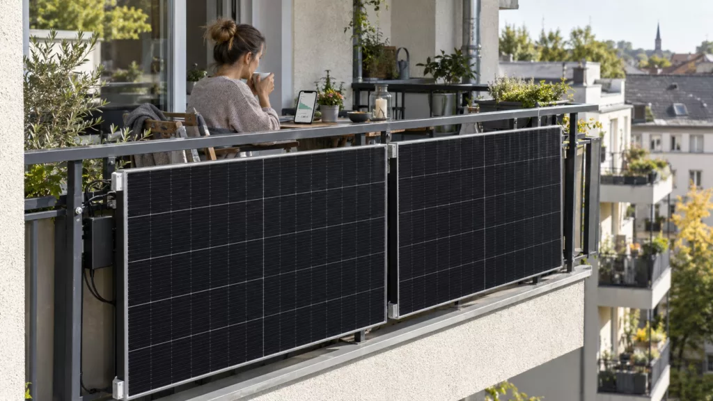 Zwei Solarmodule hängen an einem Stadtbalkon, auf dem Balkon sitzt eine Frau mit dem Rücken zum Betrachter, sie blickt auf ein Smartphone.