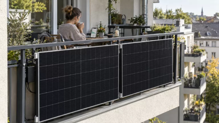 Zwei Solarmodule hängen an einem Stadtbalkon, auf dem Balkon sitzt eine Frau mit dem Rücken zum Betrachter, sie blickt auf ein Smartphone.