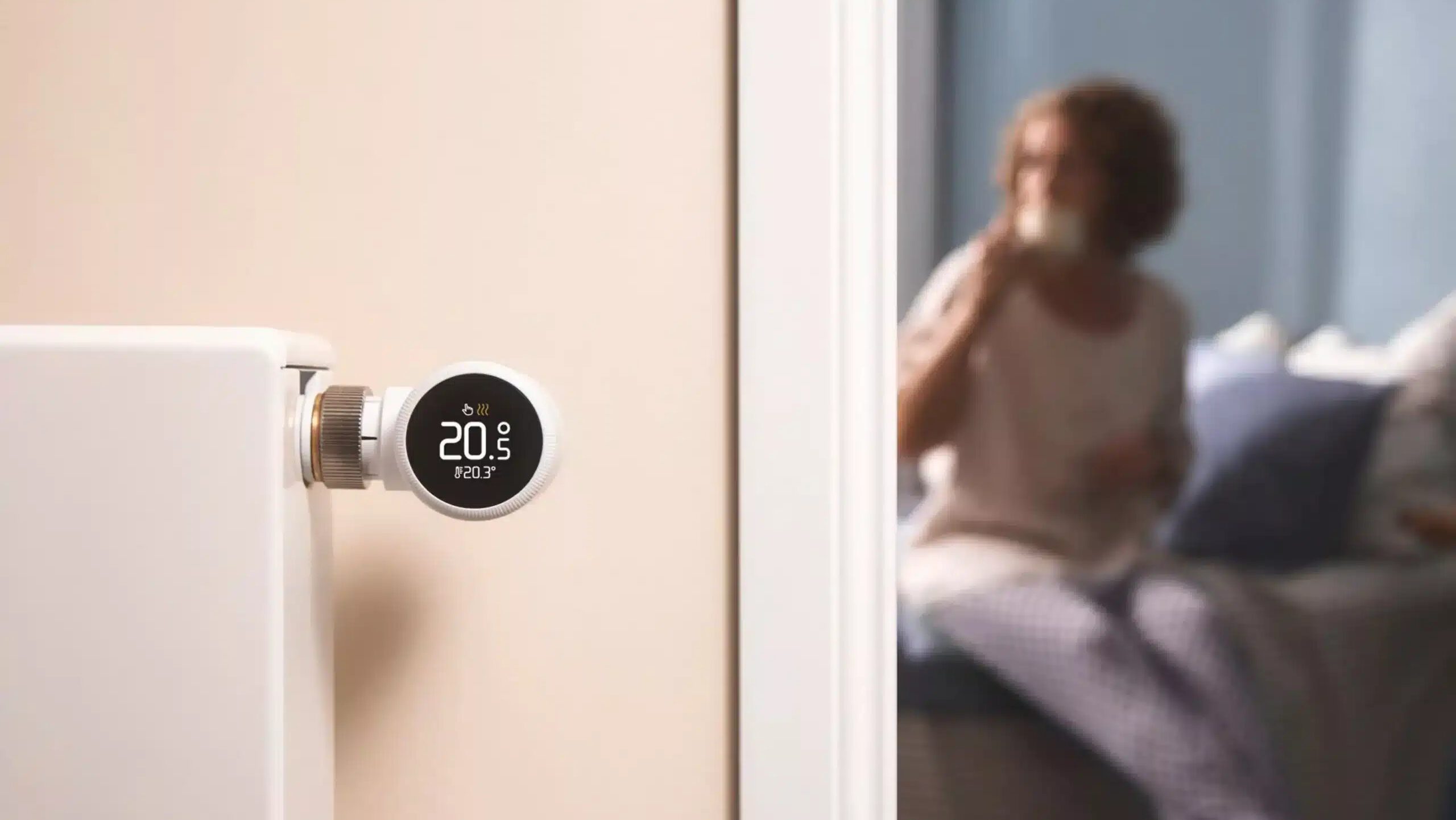Links weißer Heizkörper mit digitalem schwarzem Thermostat vor beiger Wand mit weißem Türrahmen, verschwommener Blick ins Zimmer, wo eine Frau mit Tasse auf Bettkante sitzt