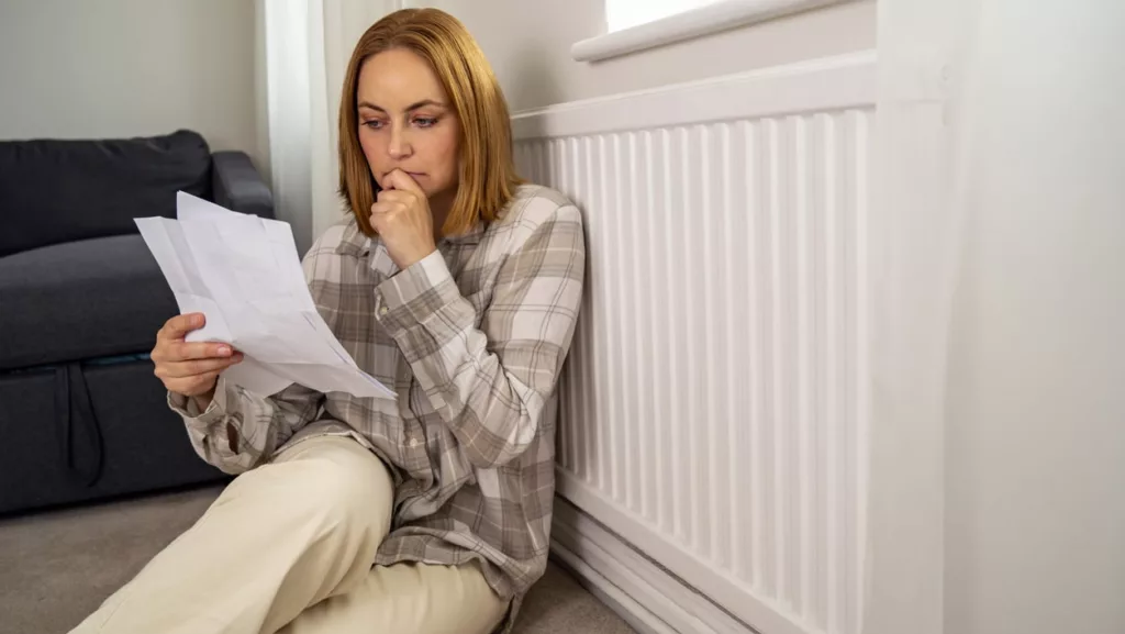 Besorgte Frau, die Papierrechnungen neben dem Heizungsthermostat hält und die Heizkosten in die Höhe treibt