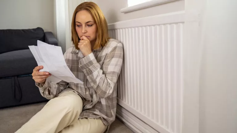 Besorgte Frau, die Papierrechnungen neben dem Heizungsthermostat hält und die Heizkosten in die Höhe treibt