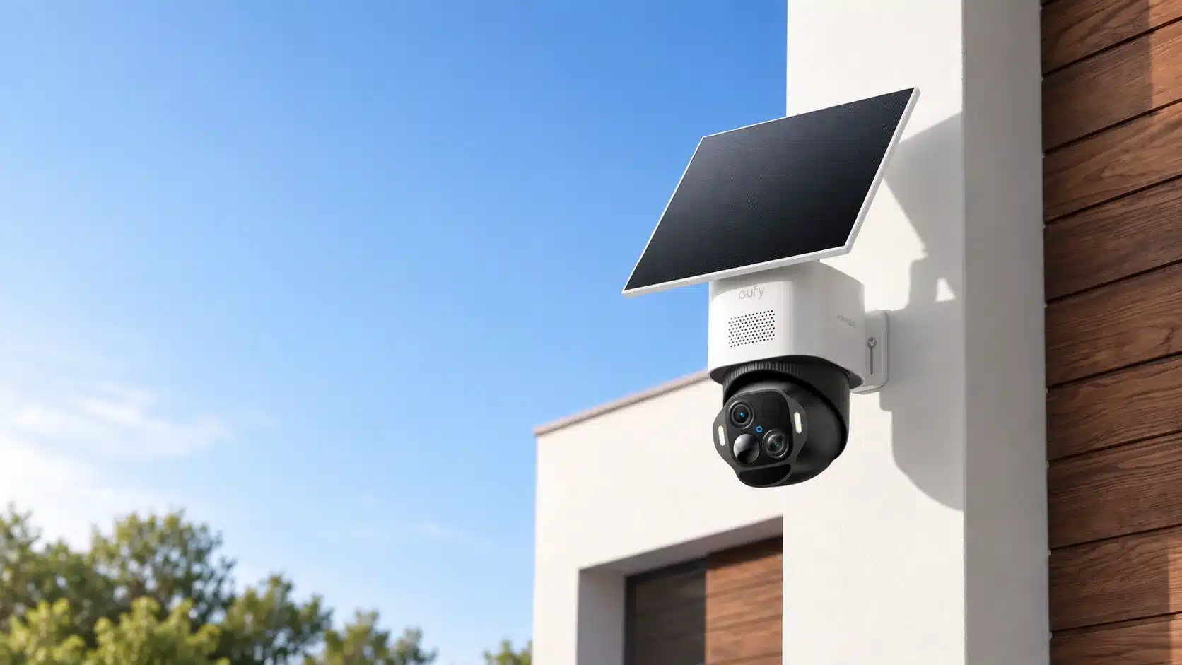 Rechts im Bild weiße Hausecke mit weiß schwarzer Kamera mit schwarzem Solarpanel oben drauf, links blauer Himmel und ein paar Baumkronen