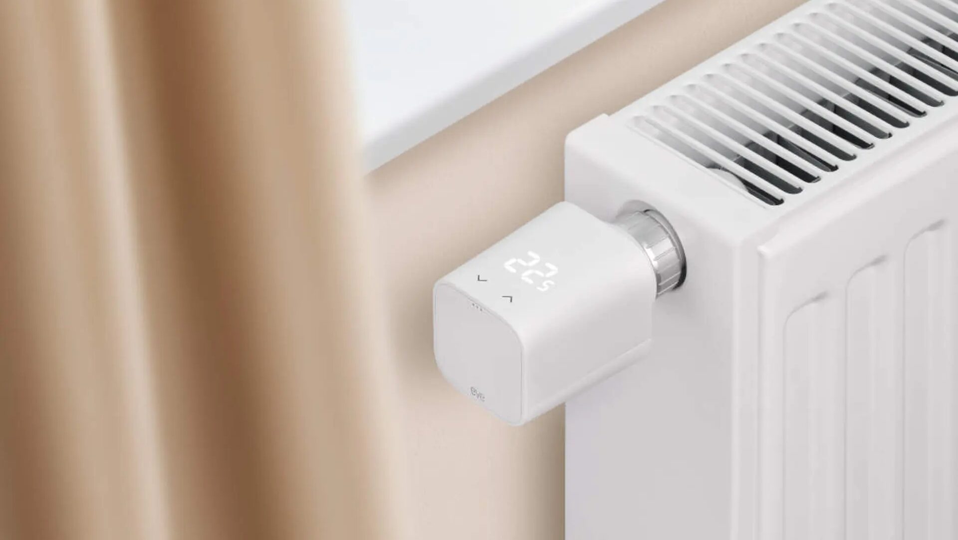 Blick auf Ende von weißer Heizung mit weißem digitalem Thermostat vor beiger Wand unter weißer Fensterbank mit beigem Vorhang links an der Seite