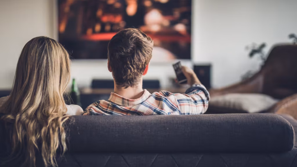 Back view of a couple watching TV on sofa in the living room. Man is changing channels.